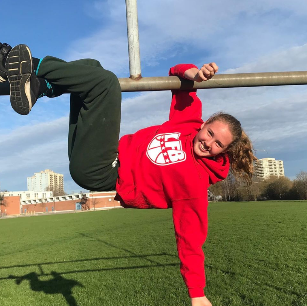 A girl in a red sweatshirt smiles while using one hand and legs to hang from a horizontal support